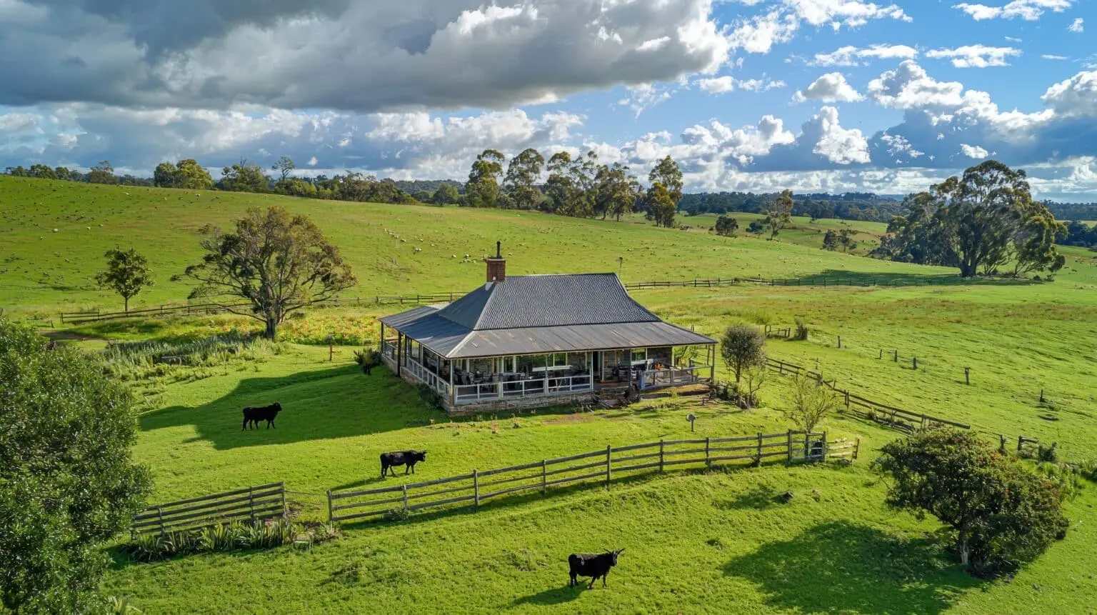 Aerial view of a countryside barndominium surrounded by fenced green pastures, grazing cattle, rolling hills, and dramatic cloudy skies.