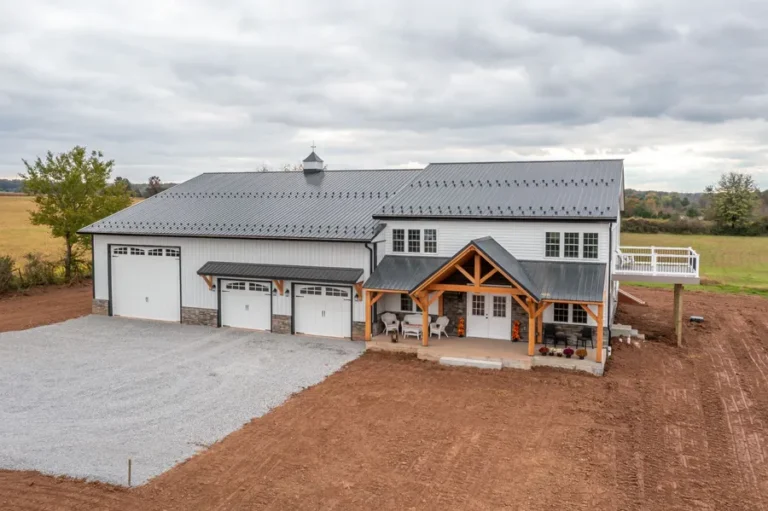 Large white barndominium with black metal roof, multiple garage bays, timber-framed front porch, and gravel driveway on a spacious rural property under cloudy skies.