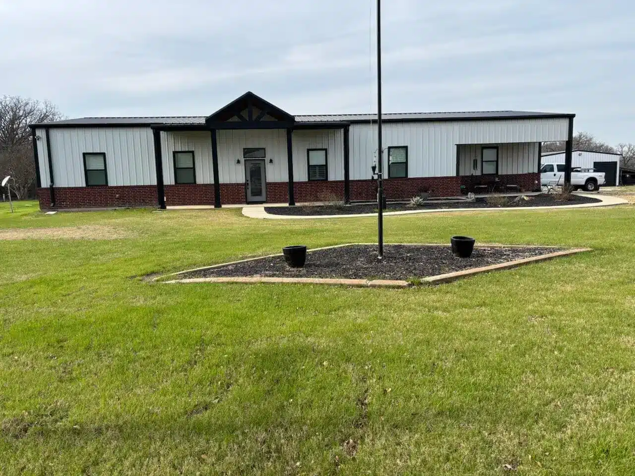 Single-story white barndominium with black trim, covered front porch, metal roof, and landscaped lawn on a spacious rural property.